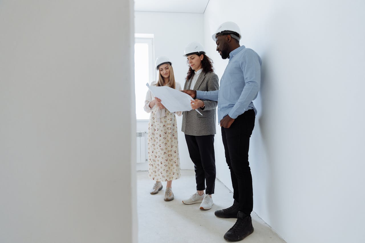 Three architects in hard hats review blueprints in a bright, empty white room.